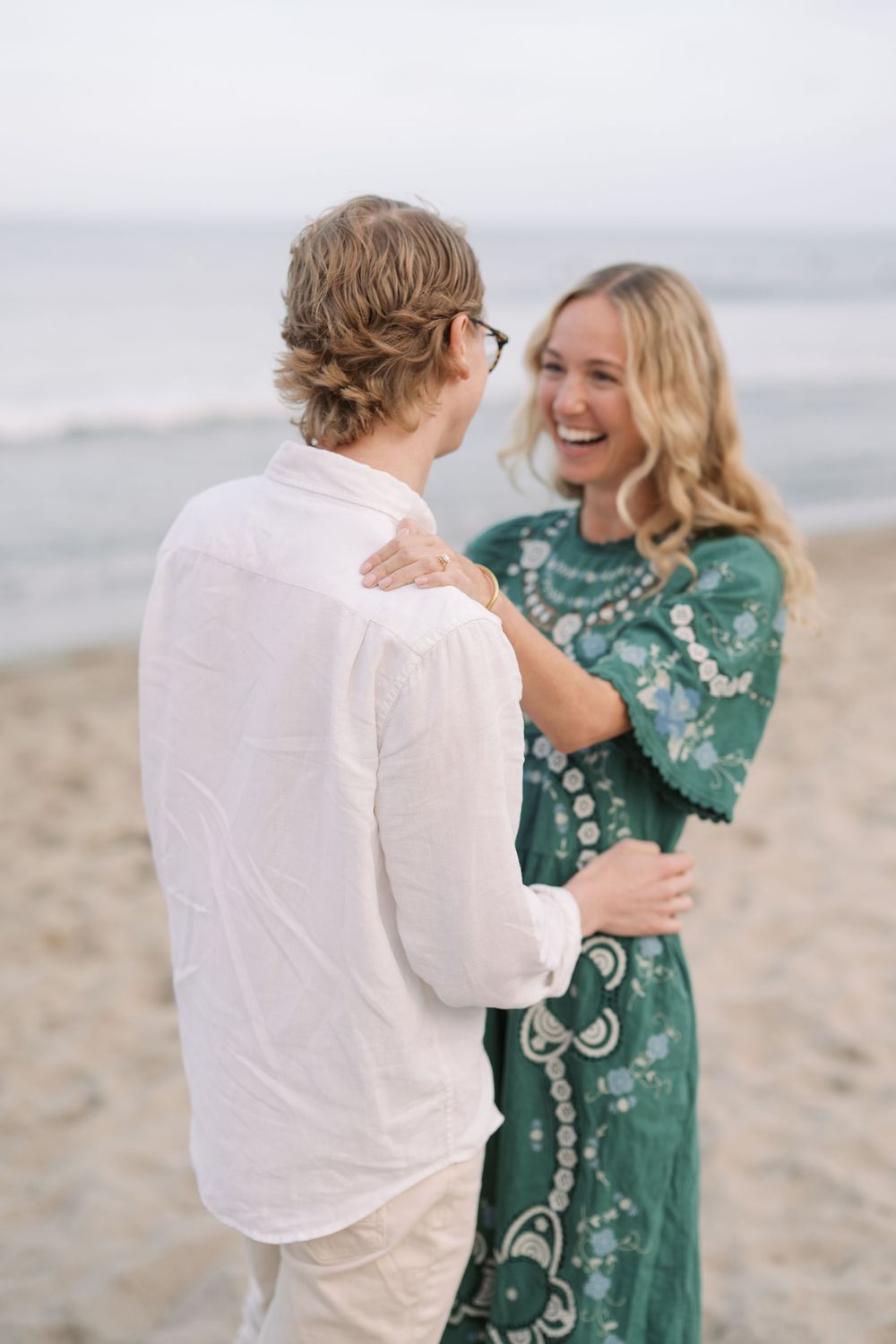 A laughing blonde woman in a green lace dress embraces a man in a white linen shirt on a sandy beach.