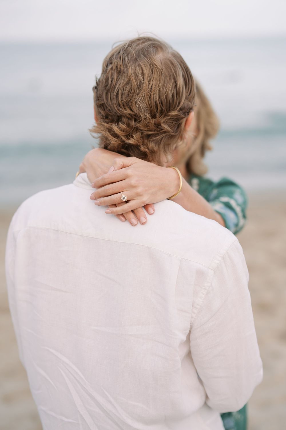 Woman with braided hair places hand with engagement ring on man's shoulder at beach.