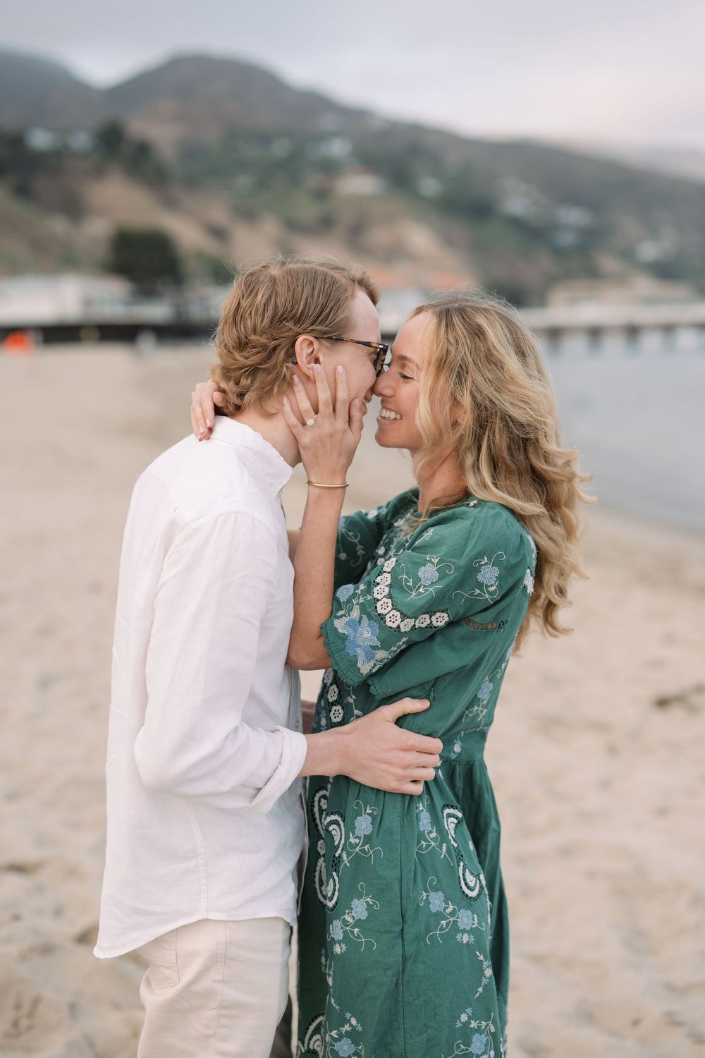 Couple sharing a romantic moment on a sandy beach with mountains in the background.