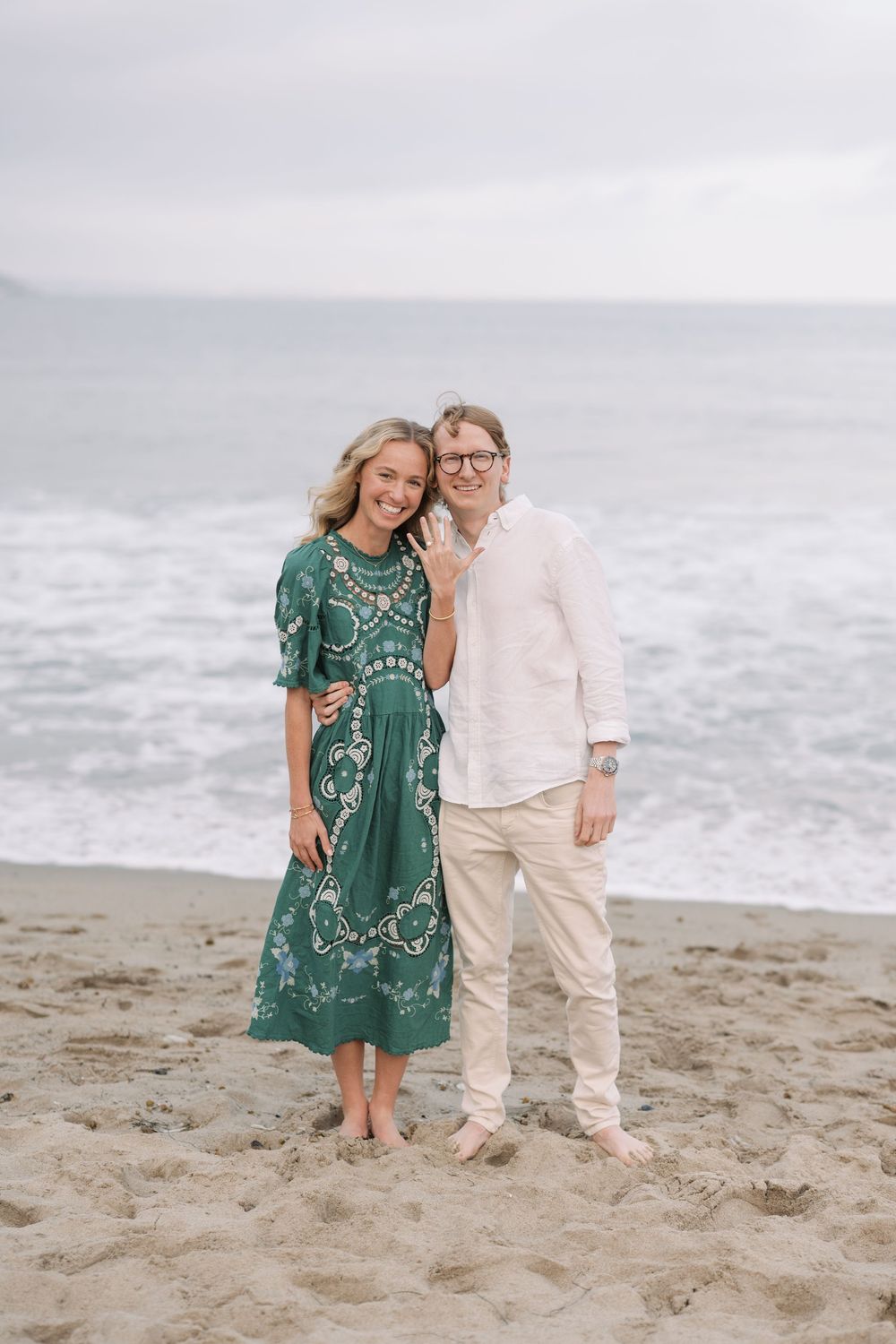 Smiling couple posing barefoot on a sandy beach, woman in green floral dress, man in white linen shirt, ocean waves behind.