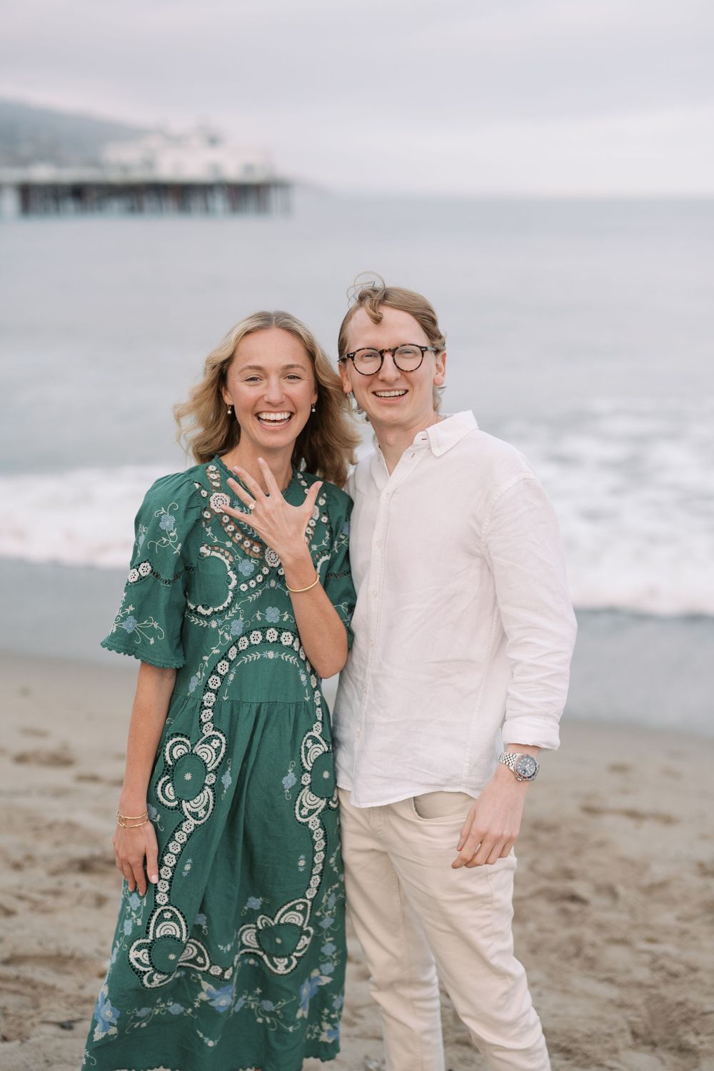 Happy couple on beach showing engagement ring, woman in green dress, man in white shirt, ocean pier backdrop.