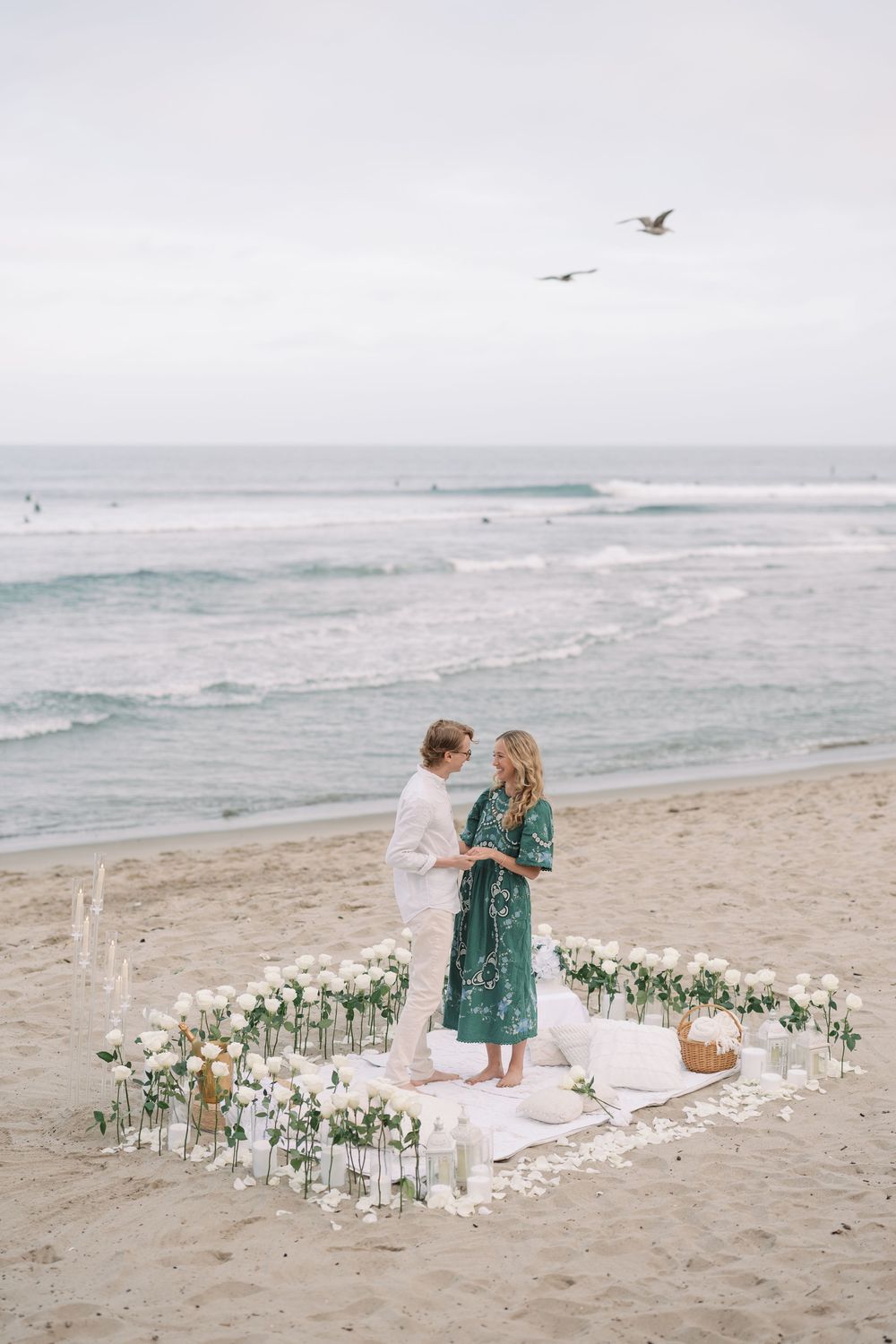 Couple stands inside a heart of white roses on a beach with seagulls flying overhead during a romantic proposal.