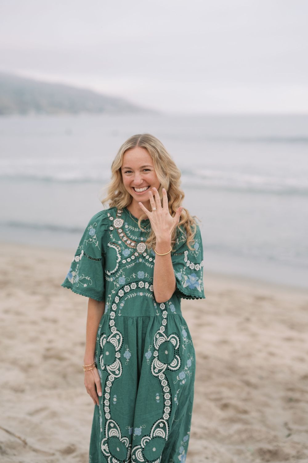 Smiling blonde woman in green boho dress showing engagement ring on beach.