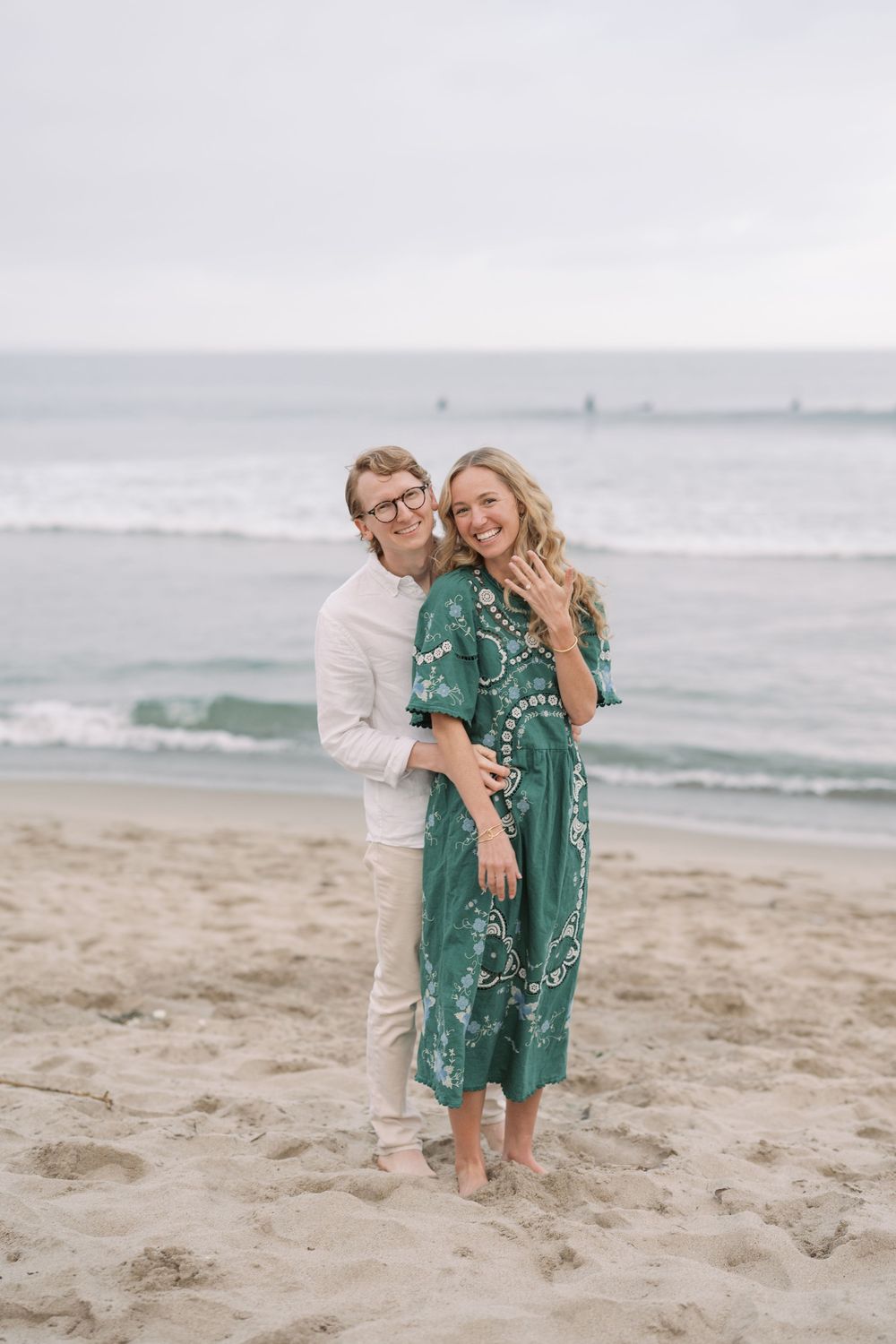 Happy couple embracing on sandy beach, woman in green floral dress showing engagement ring, ocean waves in background.