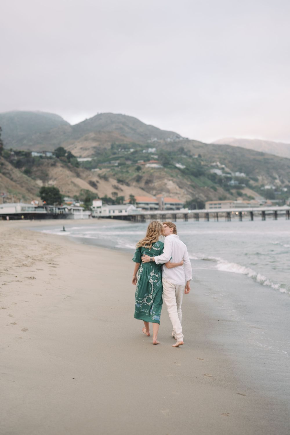 Couple walking barefoot on a sandy Malibu beach, woman in green dress, man in white, mountains in background.