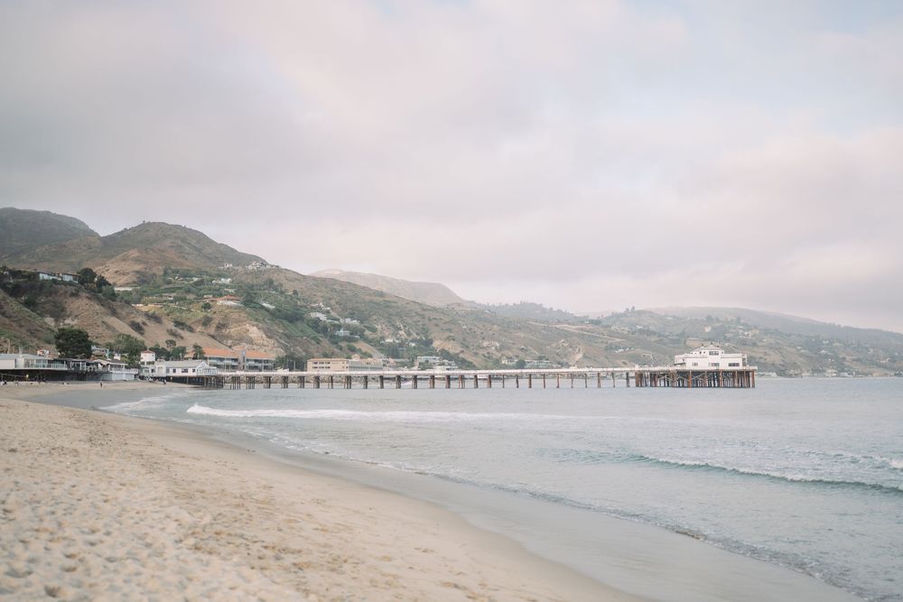 Sandy beach with a long pier extending into calm ocean waters, backed by green hills under an overcast sky in Malibu.