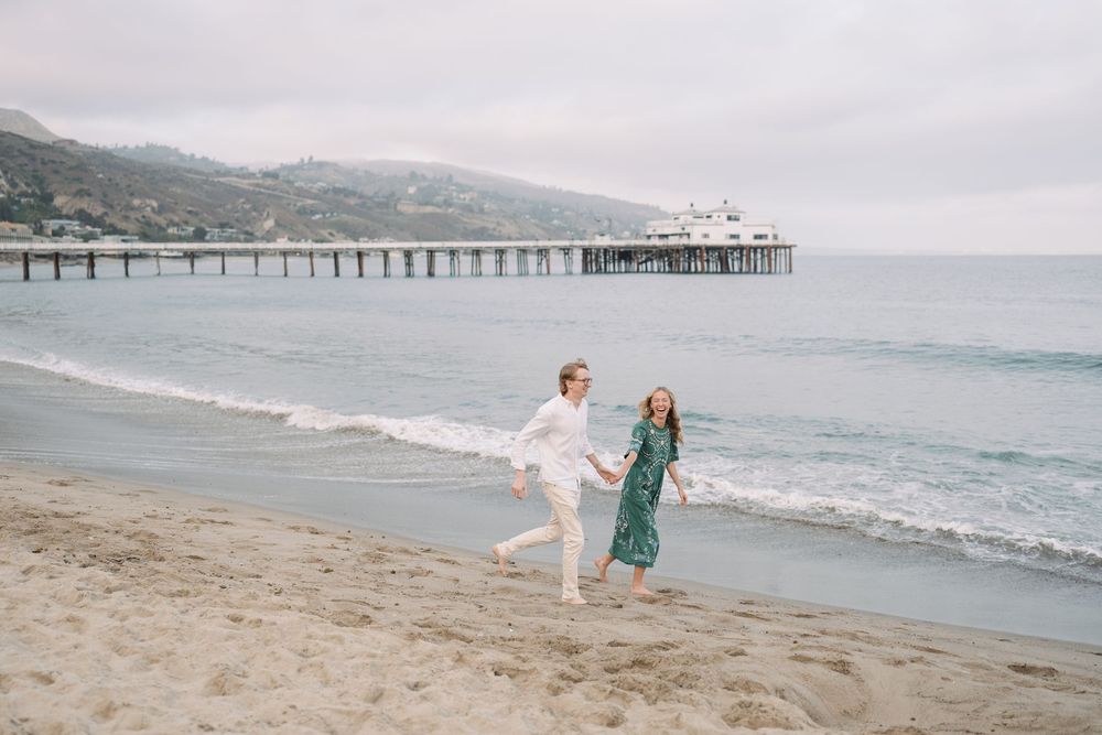 Couple holding hands running on Malibu beach with pier and ocean waves in background on cloudy day.