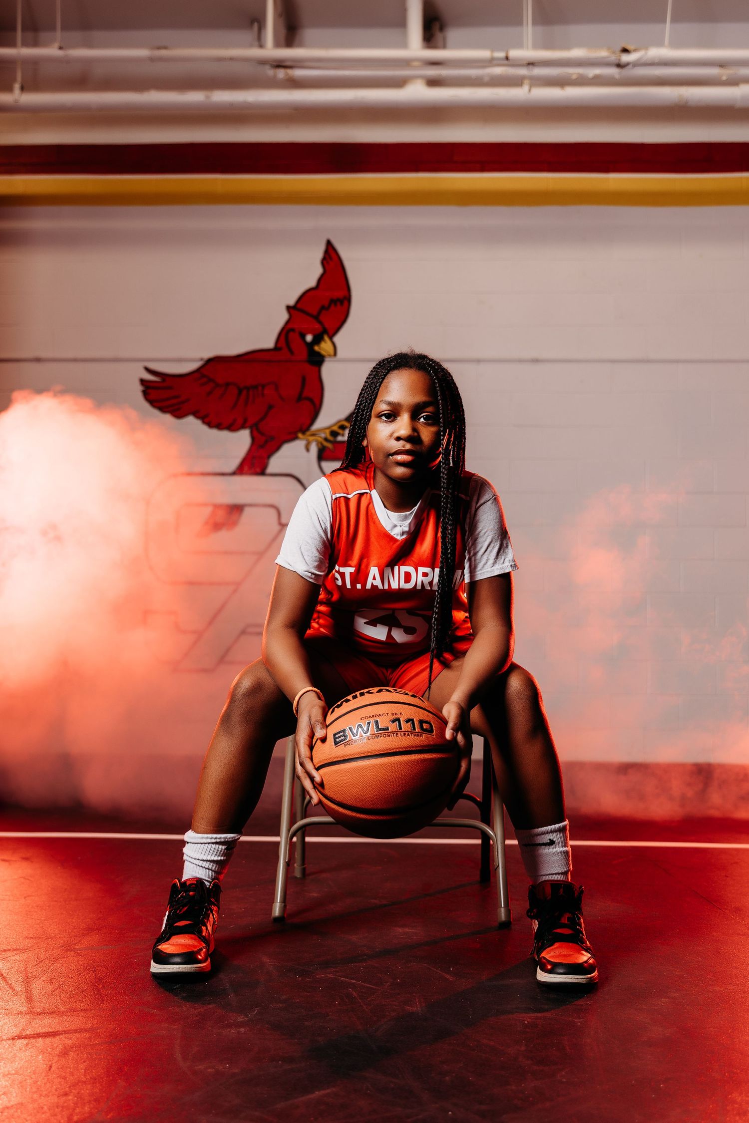 Female basketball player in St. Andrew Cardinals orange jersey sits holding a basketball on a gym floor with red dramatic lighting.