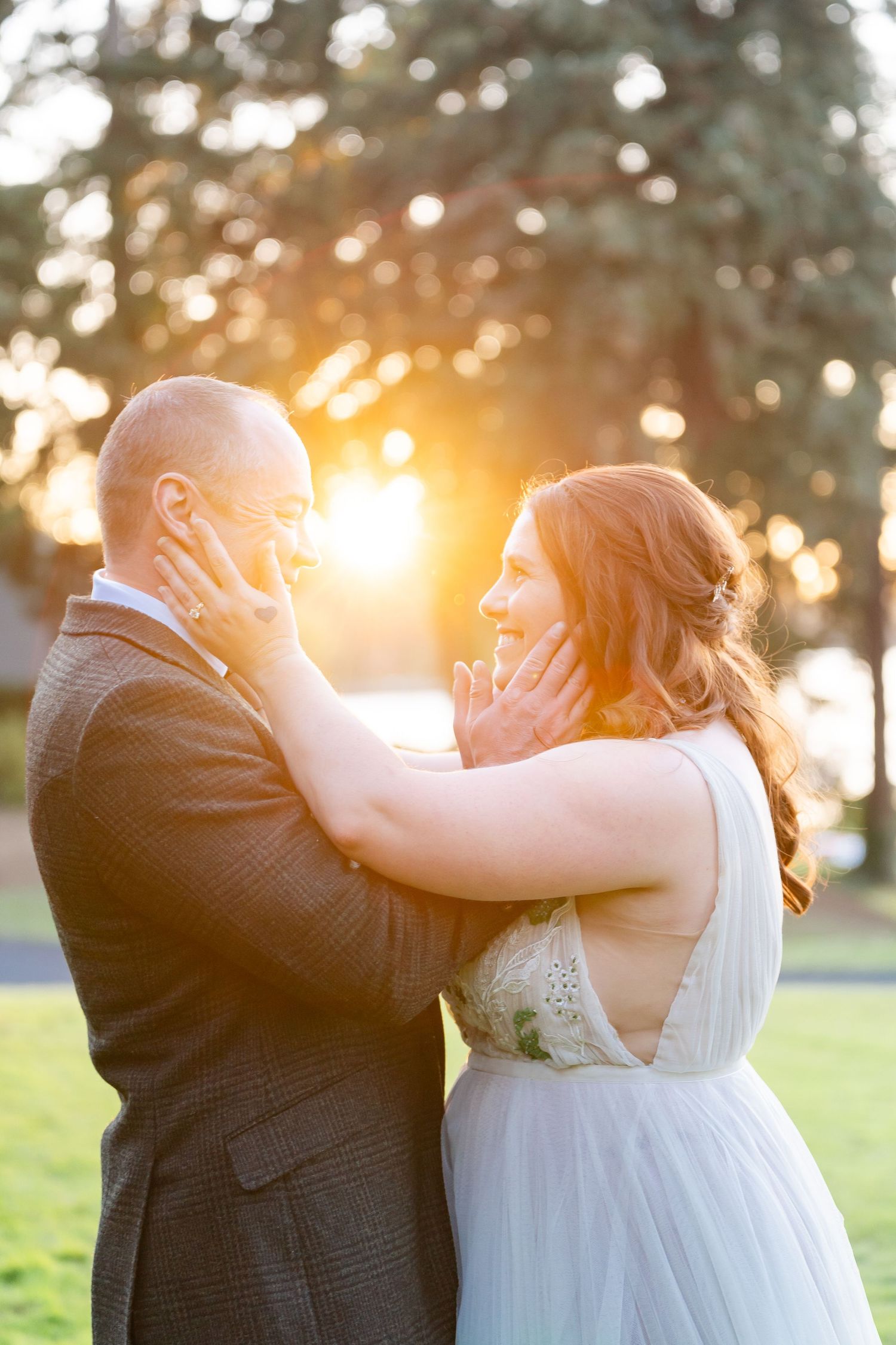 Bride and groom share a tender moment at golden hour sunset, with warm bokeh light glowing between the trees behind them.