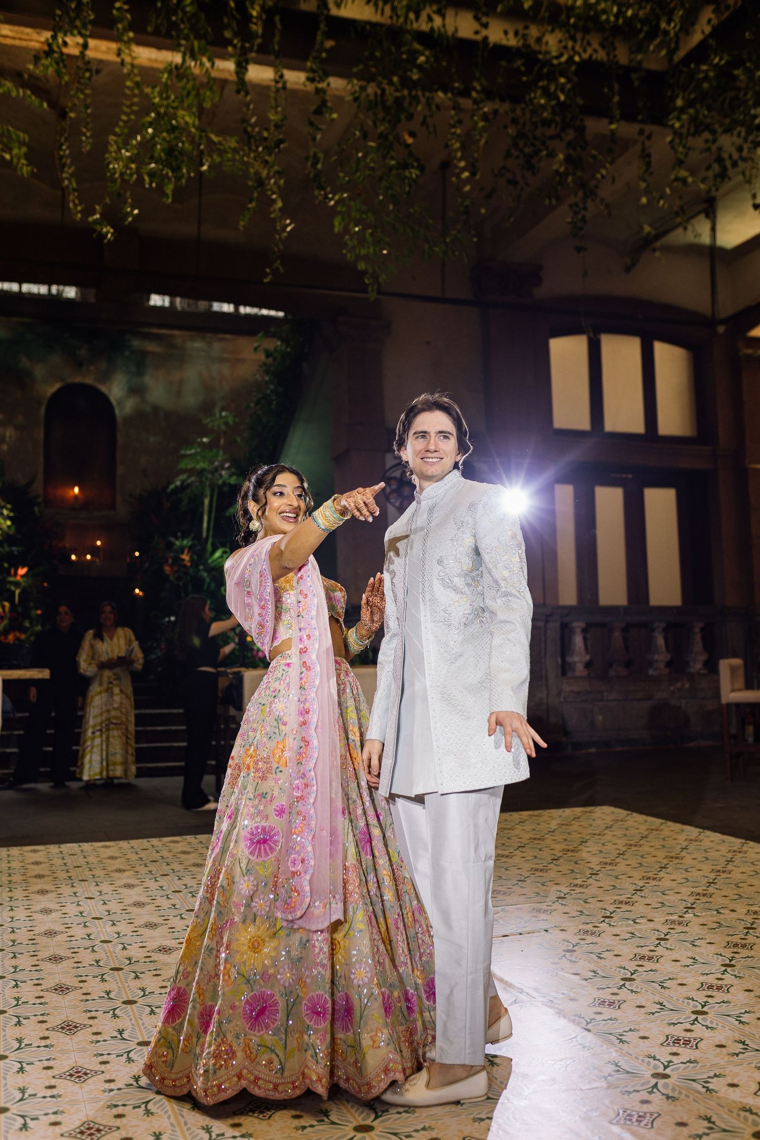 Bride in pink floral lehenga and groom in white sherwani share first dance at elegant Indian wedding reception.