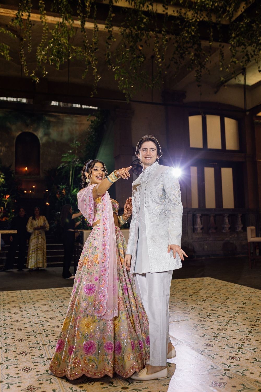 Bride in pink floral lehenga and groom in white sherwani share first dance at elegant Indian wedding reception.