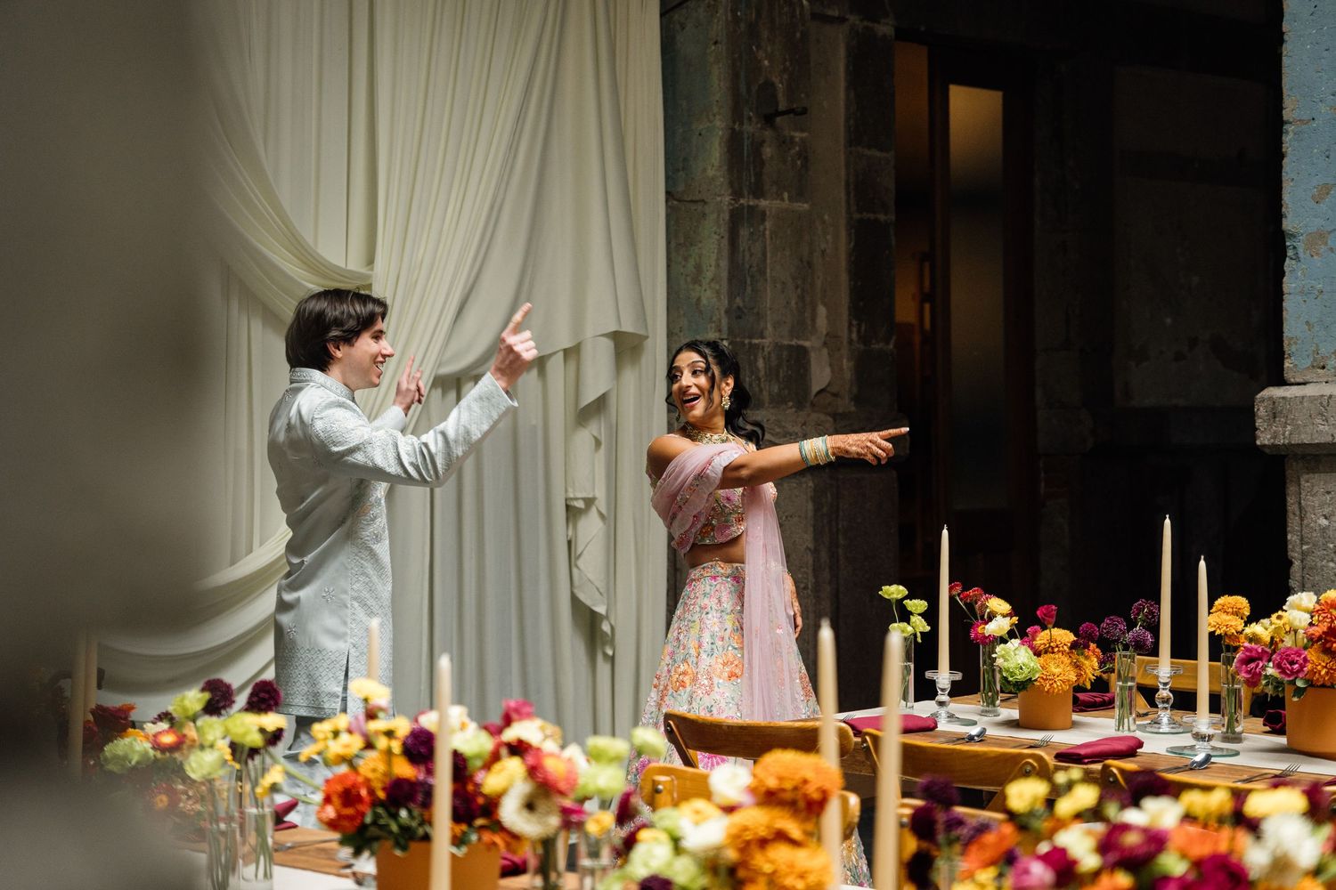 Couple gesturing excitedly at colorful floral wedding tablescape with candles in a draped venue.