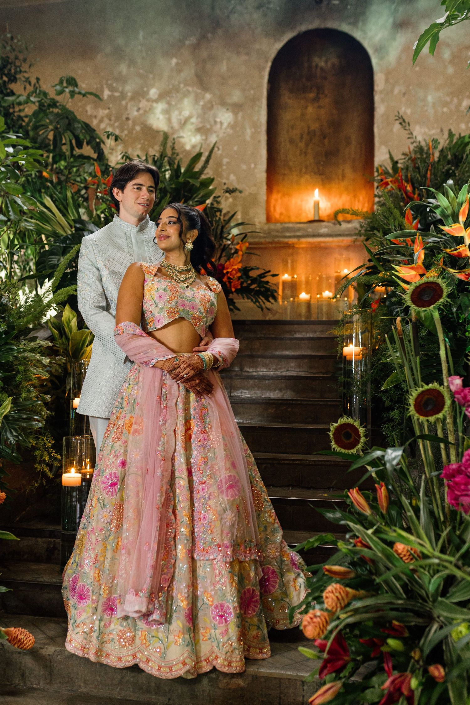 Couple in traditional Indian wedding attire surrounded by lush greenery and candles in an elegant floral setting.