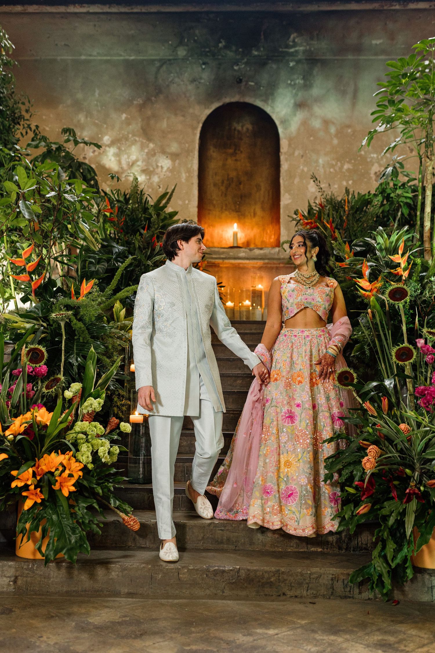 Couple in traditional Indian wedding attire holding hands among tropical flowers and candlelit stone arch backdrop.