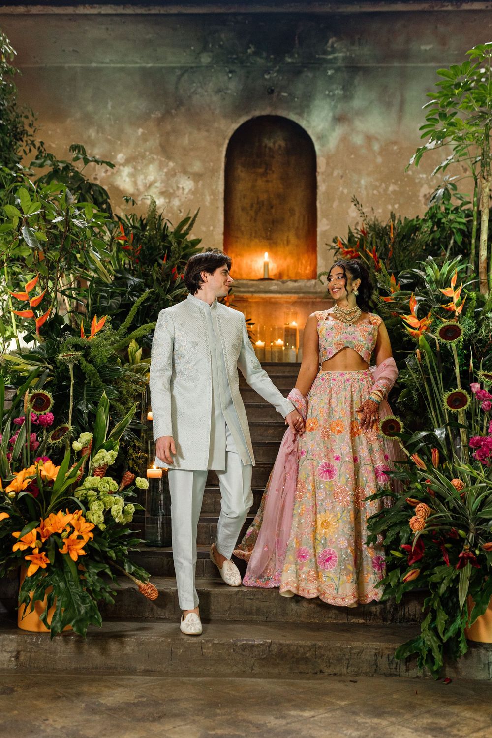Couple in traditional Indian wedding attire holding hands among tropical flowers and candlelit stone arch backdrop.