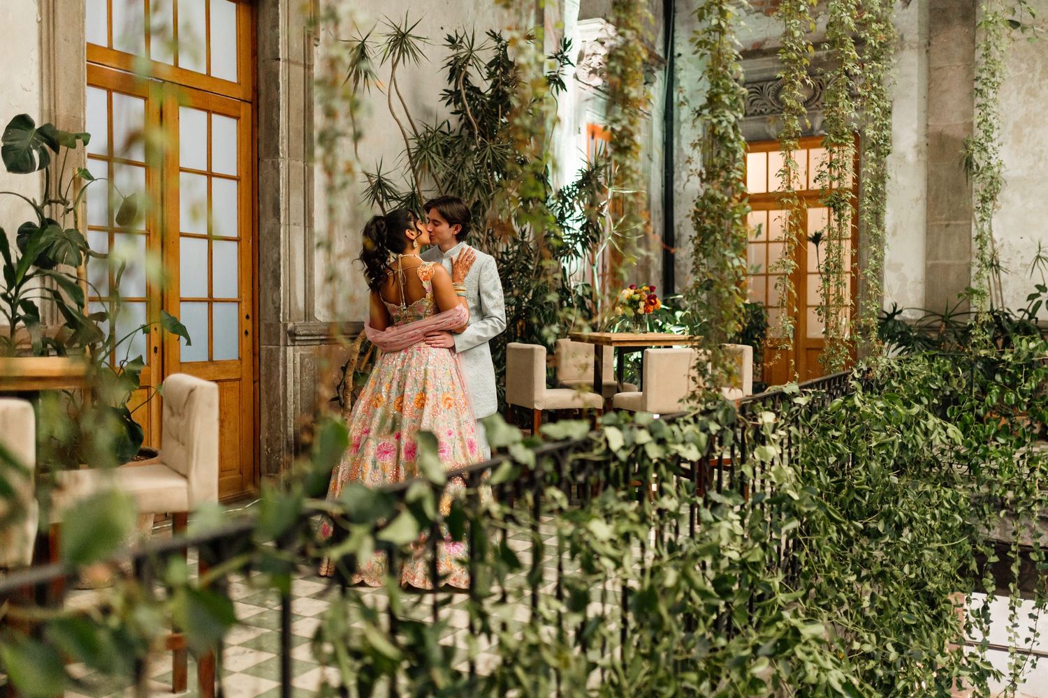 Couple sharing a romantic kiss in a lush green courtyard with ivy-covered walls and elegant French doors.