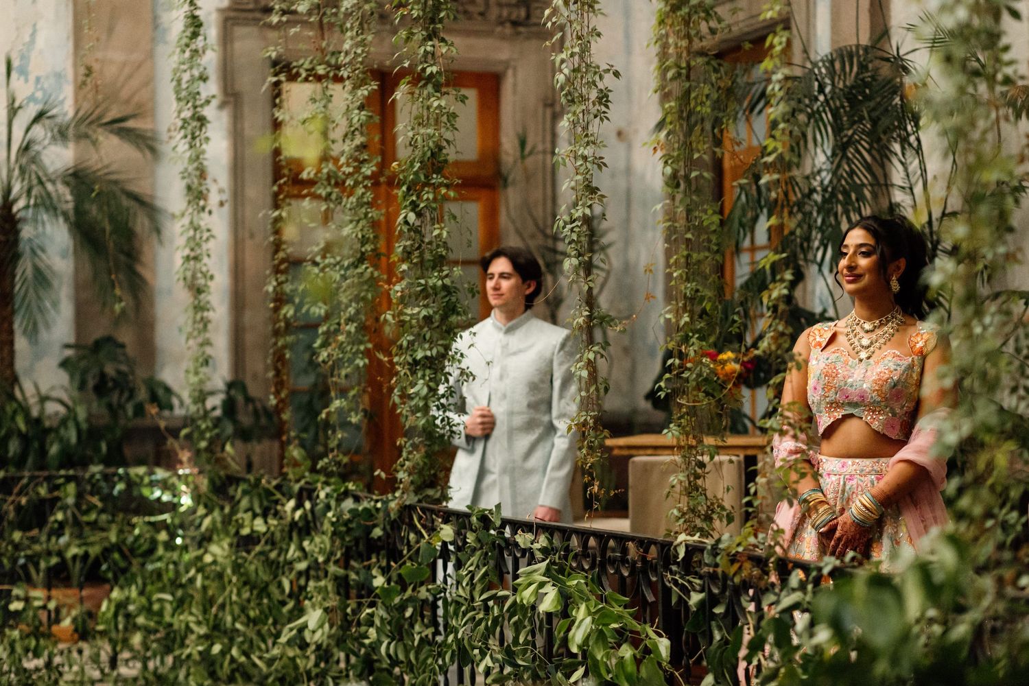 Couple in traditional Indian wedding attire standing on a lush green balcony surrounded by tropical plants.