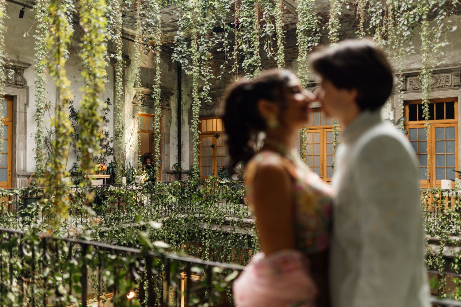 Couple sharing a romantic moment on a balcony surrounded by lush greenery and hanging vines at an elegant venue.