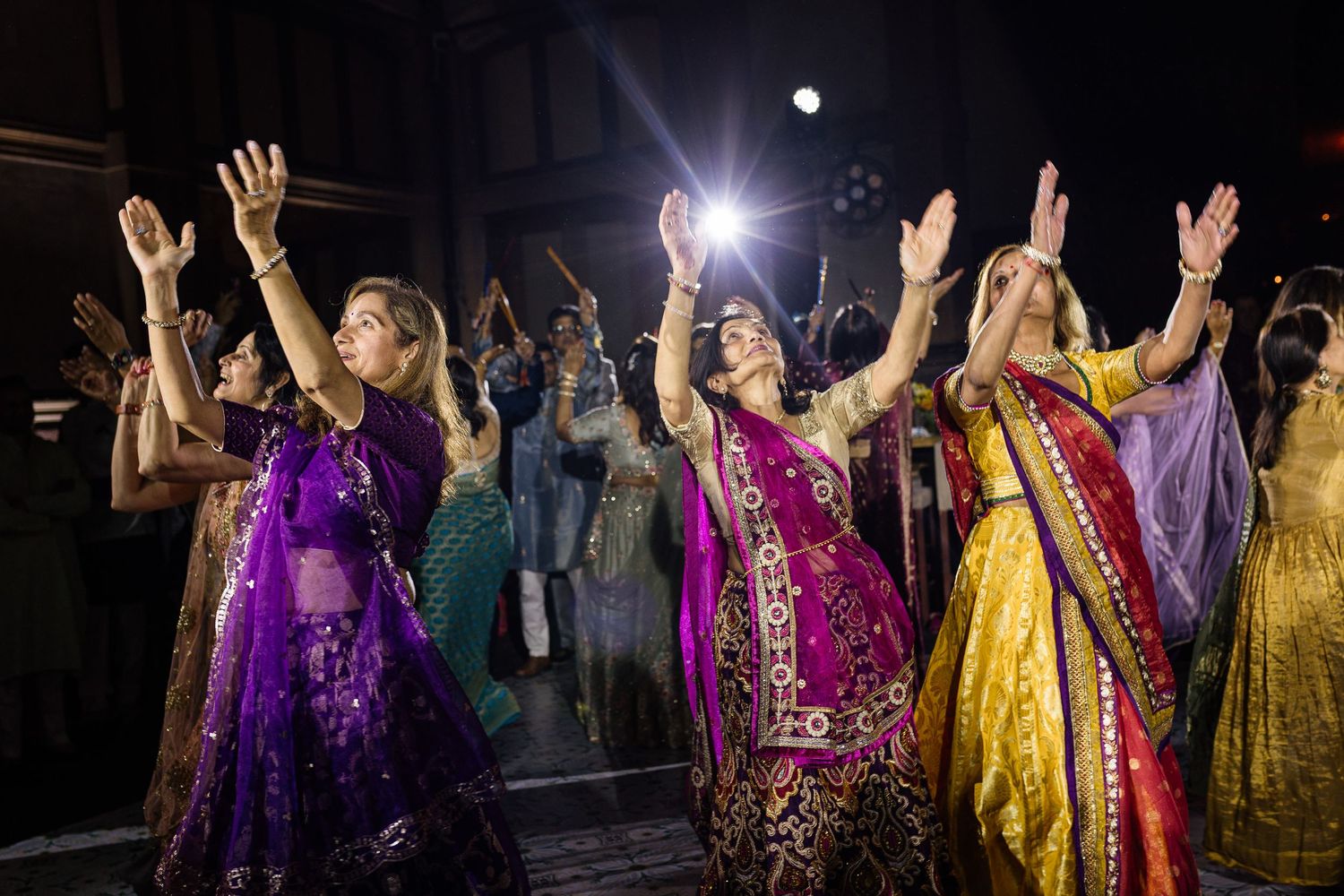 Women in vibrant traditional Indian attire dance with raised hands during a colorful cultural celebration.