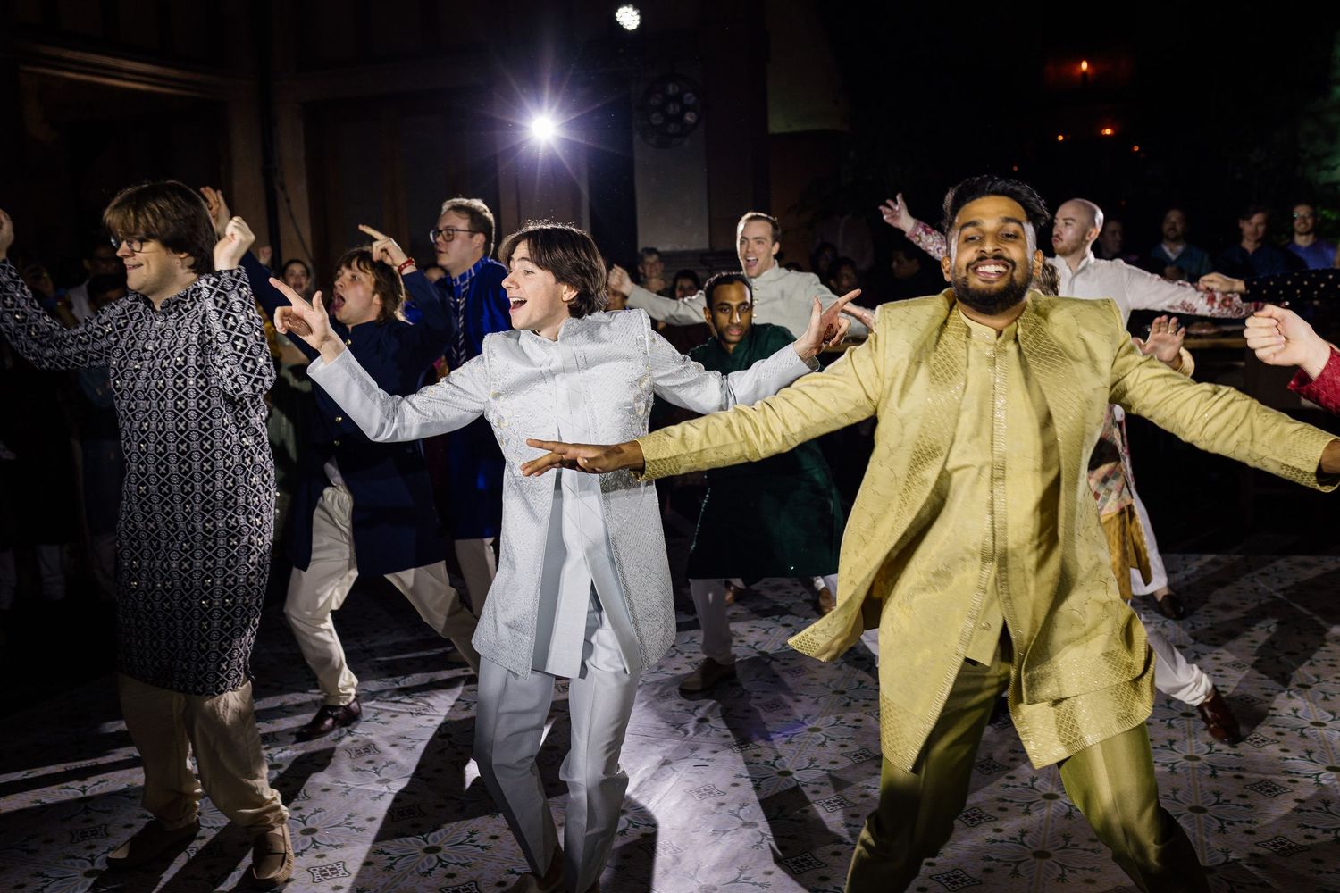 Diverse group of people dancing joyfully at a festive celebration, with a man in a gold outfit leading.