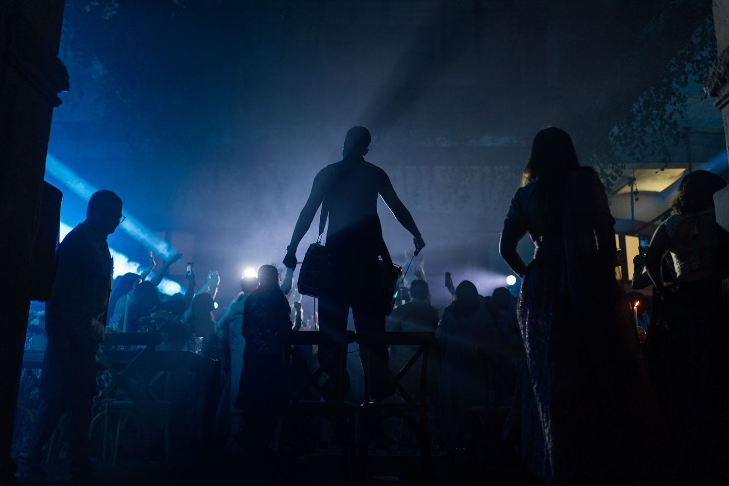 Silhouetted figures dance in a dark, smoky nightclub illuminated by dramatic blue stage lights.