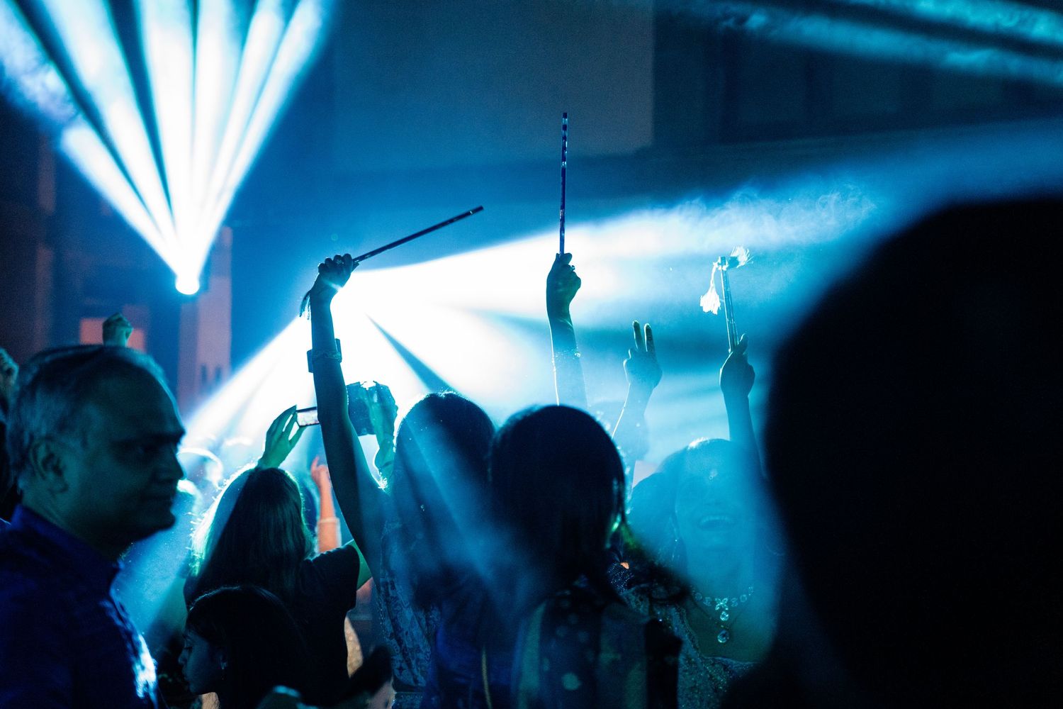 Crowd dancing under blue stage lights at a nightclub concert with raised hands and light sticks.