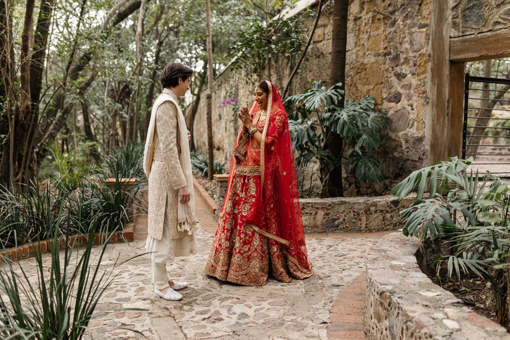 Bride in red lehenga and groom in ivory sherwani walk hand in hand on stone path in lush garden setting.