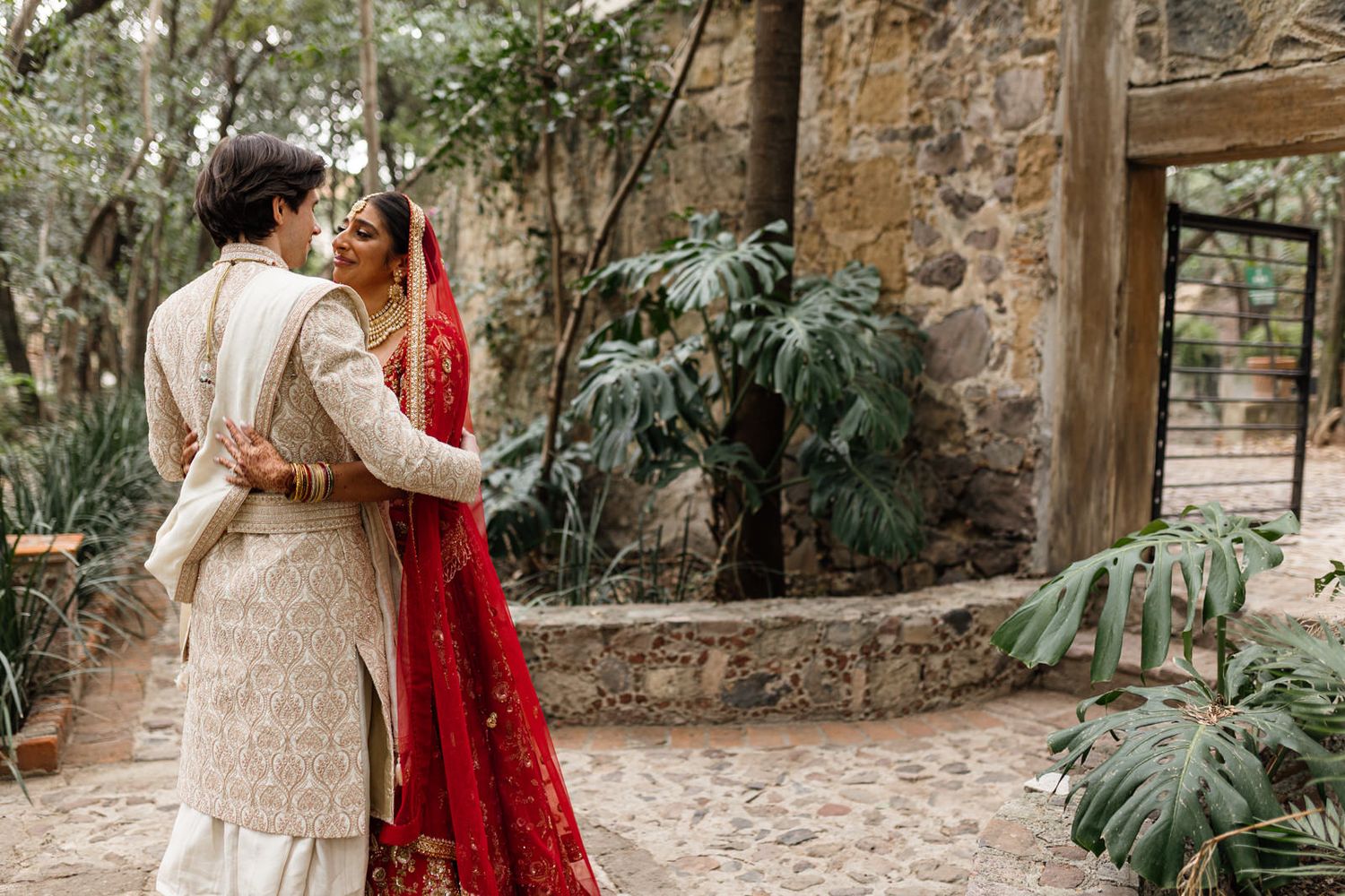 Couple in traditional Indian wedding attire embrace outdoors, bride in red saree and groom in ivory sherwani.