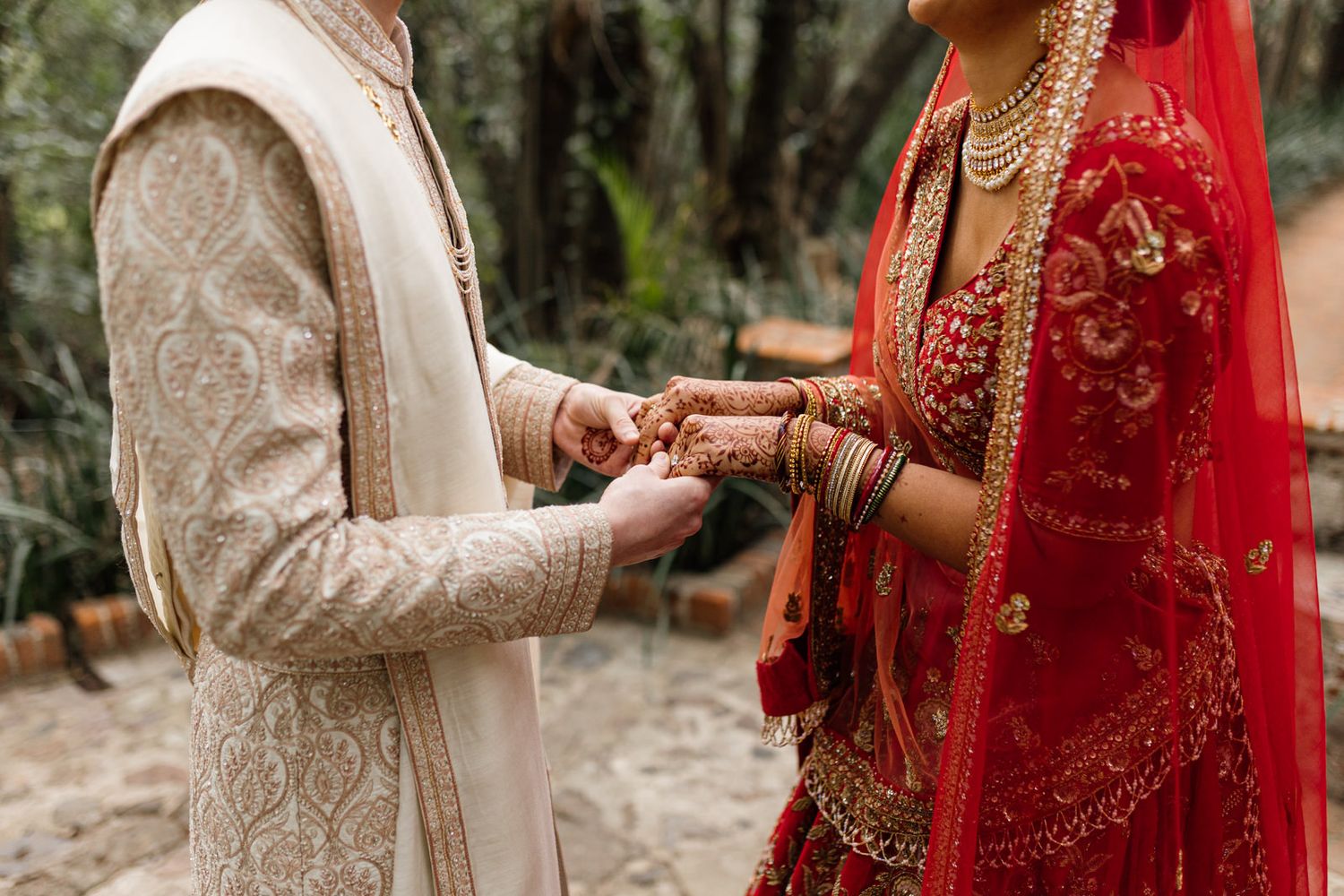 Indian bride in red lehenga and groom in beige sherwani exchanging rings during traditional Hindu wedding ceremony.