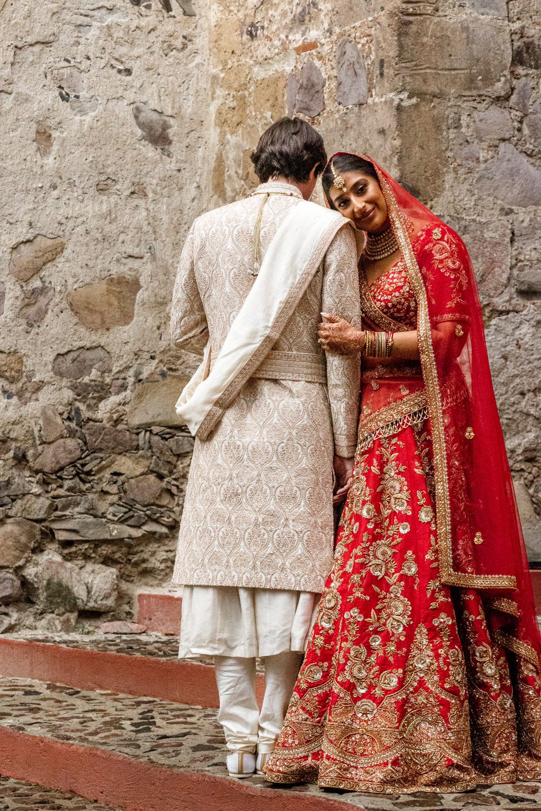 Indian bride in red lehenga and groom in beige sherwani share a tender moment at their traditional wedding.