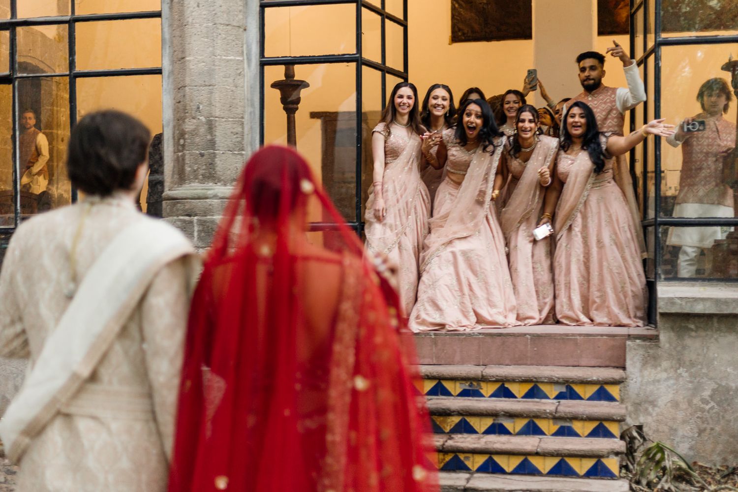 Groom in white sherwani watching bride in red dupatta walk toward bridesmaids on decorated steps at Indian wedding.