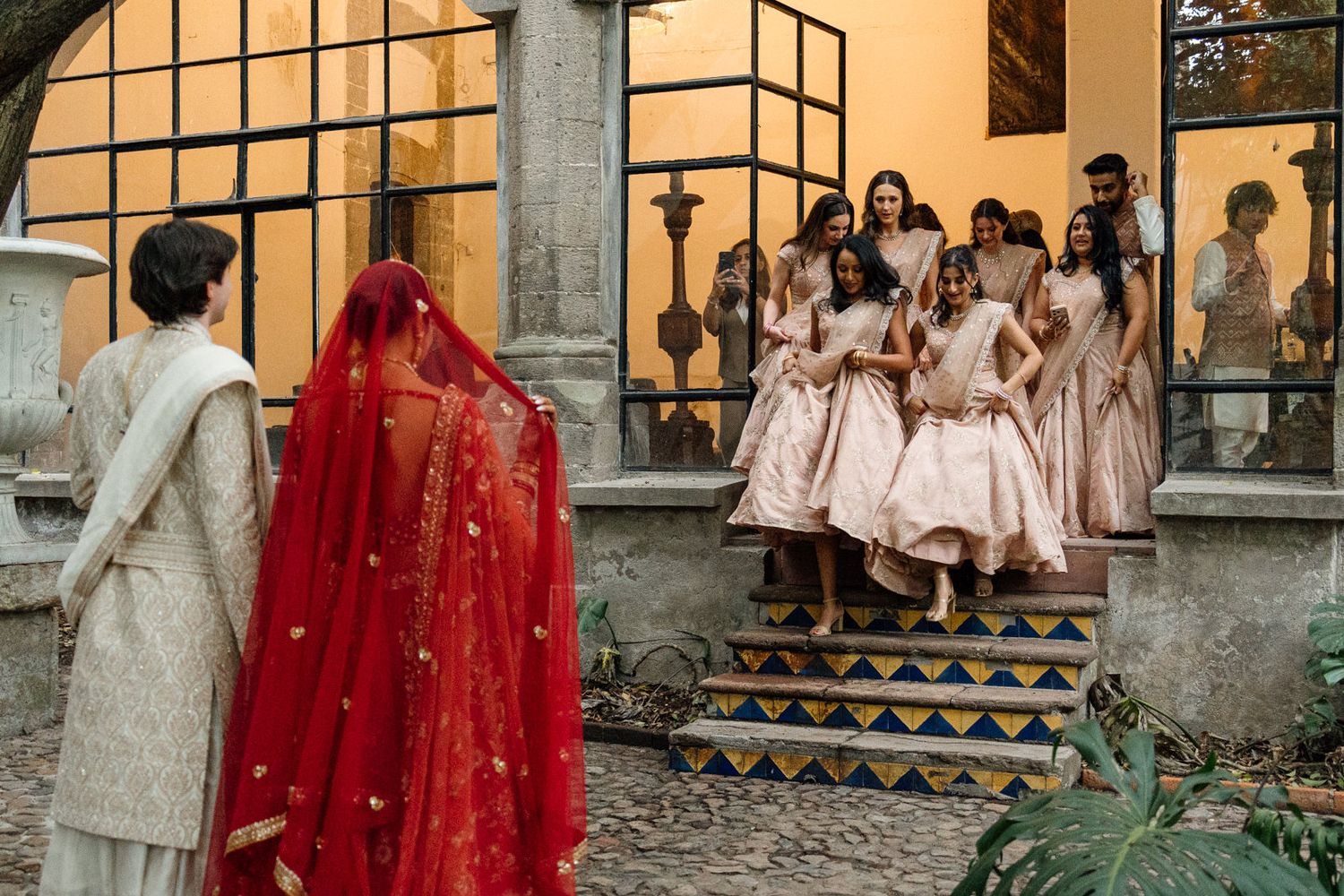 Indian bride in red veil with bridesmaids in pink dresses on ornate steps at a traditional wedding ceremony.