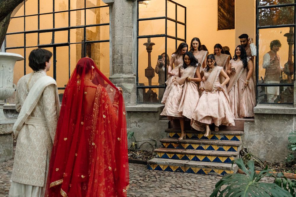 Indian bride in red veil with bridesmaids in pink dresses on ornate steps at a traditional wedding ceremony.