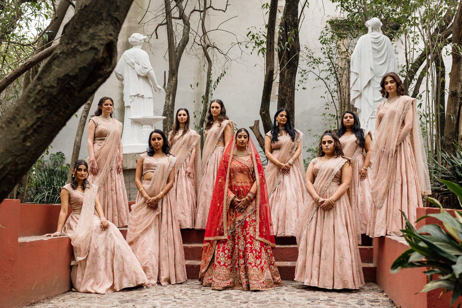 Indian bride in red lehenga surrounded by bridesmaids in pink sarees posing in a garden with white statues.