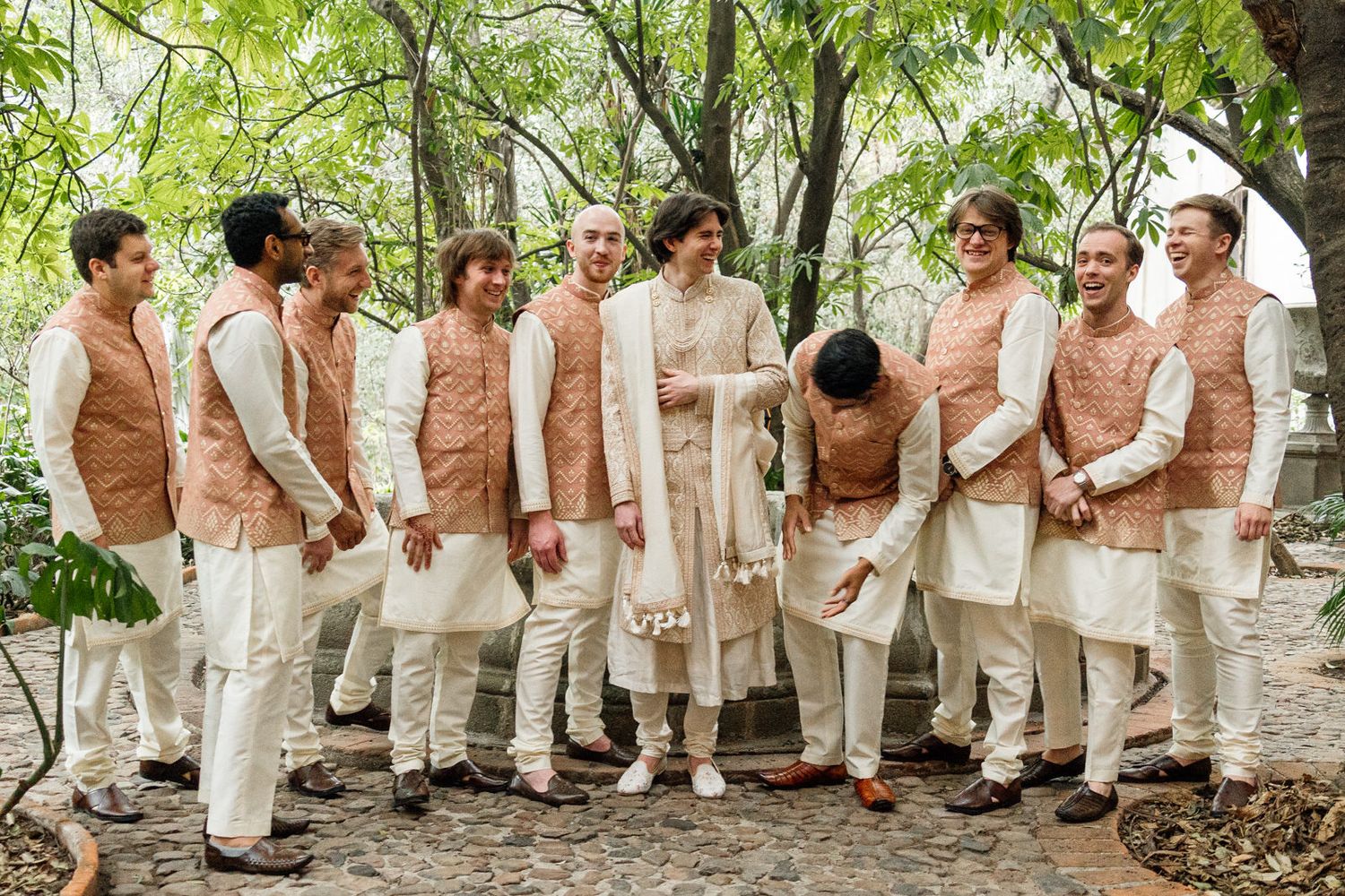 Groom and groomsmen in traditional Indian attire with cream kurtas and rose gold vests outdoors.