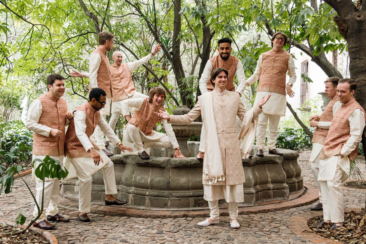 Group of men in elegant cream and peach traditional Indian attire posing around a stone fountain in a lush garden.
