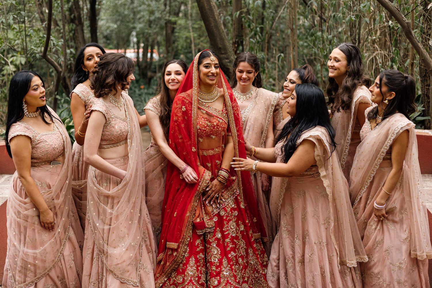 Indian bride in red lehenga surrounded by bridesmaids in blush pink outfits laughing together outdoors.