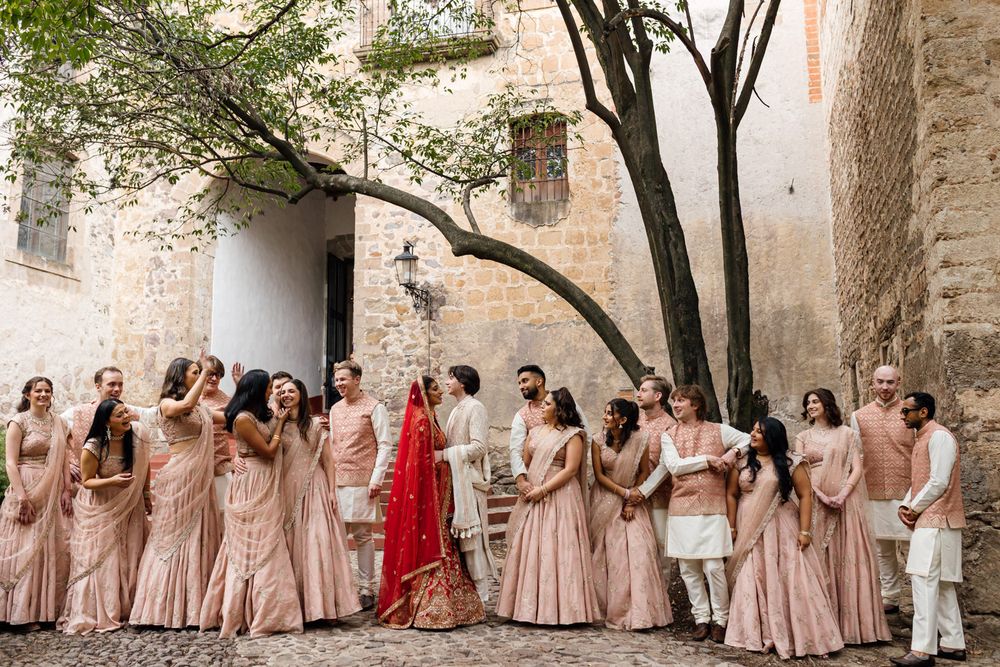 Indian wedding party in blush pink lehengas and sherwanis gathered in a rustic stone courtyard with a bride in red.
