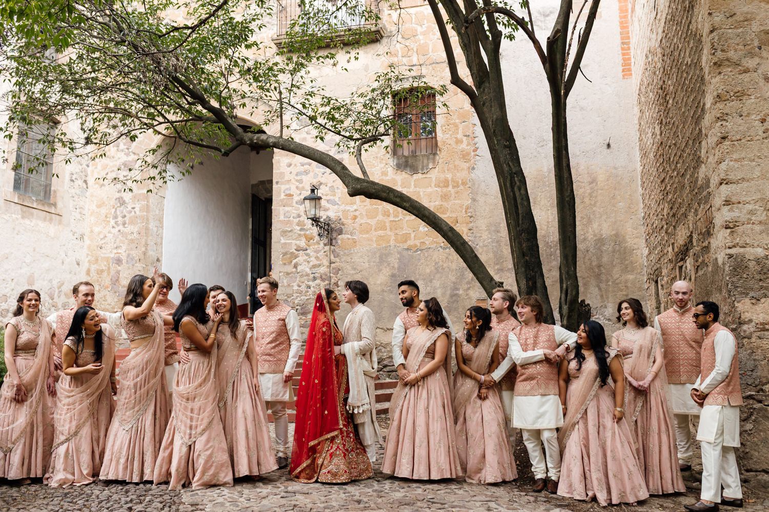 Indian wedding party in blush pink lehengas and sherwanis gathered in a rustic stone courtyard with a bride in red.