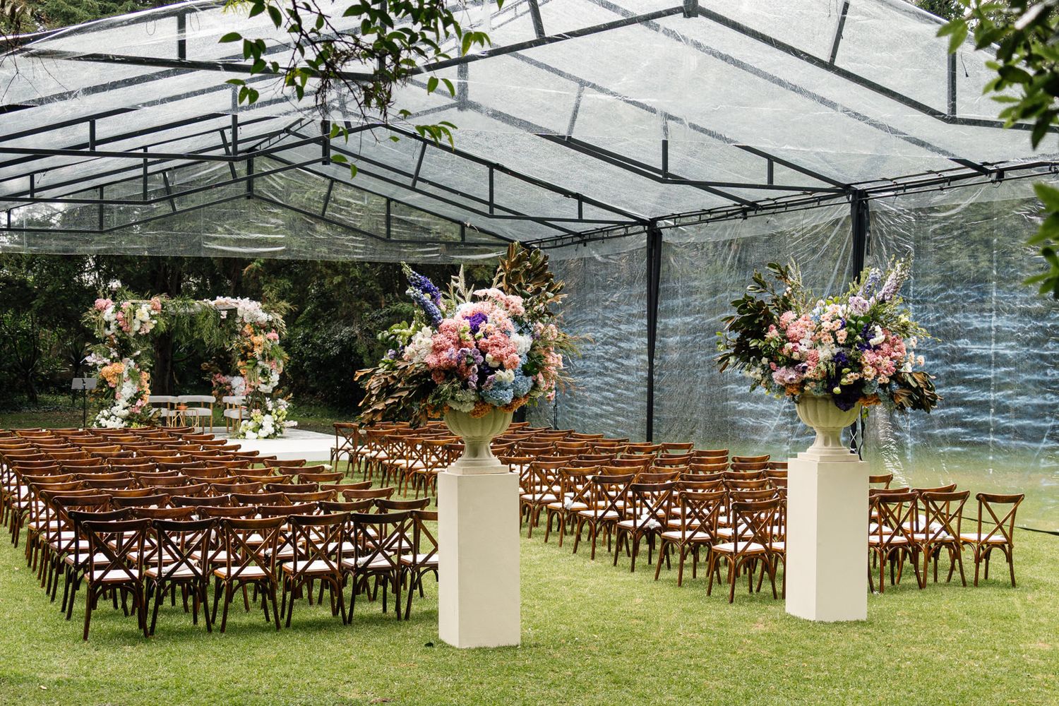 Elegant outdoor wedding ceremony setup under a clear tent with floral arrangements and wooden cross-back chairs on grass.
