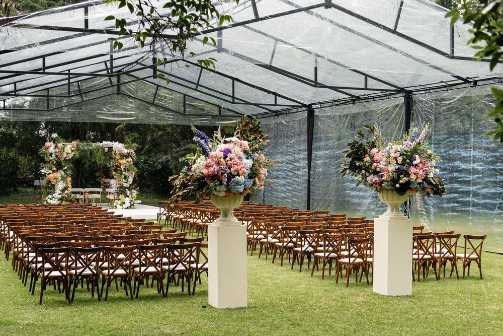 Elegant outdoor wedding ceremony setup under a clear tent with floral arrangements and wooden cross-back chairs on grass.