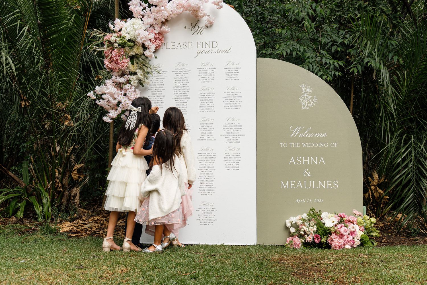 Three flower girls in white dresses view a wedding seating chart sign for Ashna and Meaulnes outdoor ceremony.