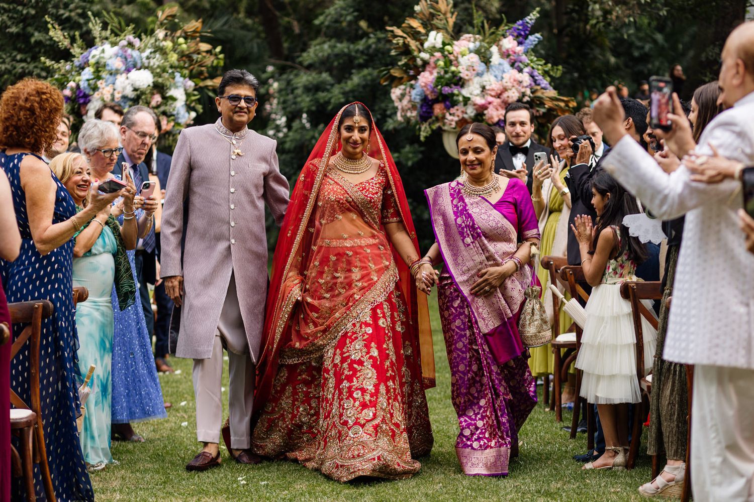 Bride in red lehenga walks down outdoor aisle with family at traditional Indian wedding ceremony.