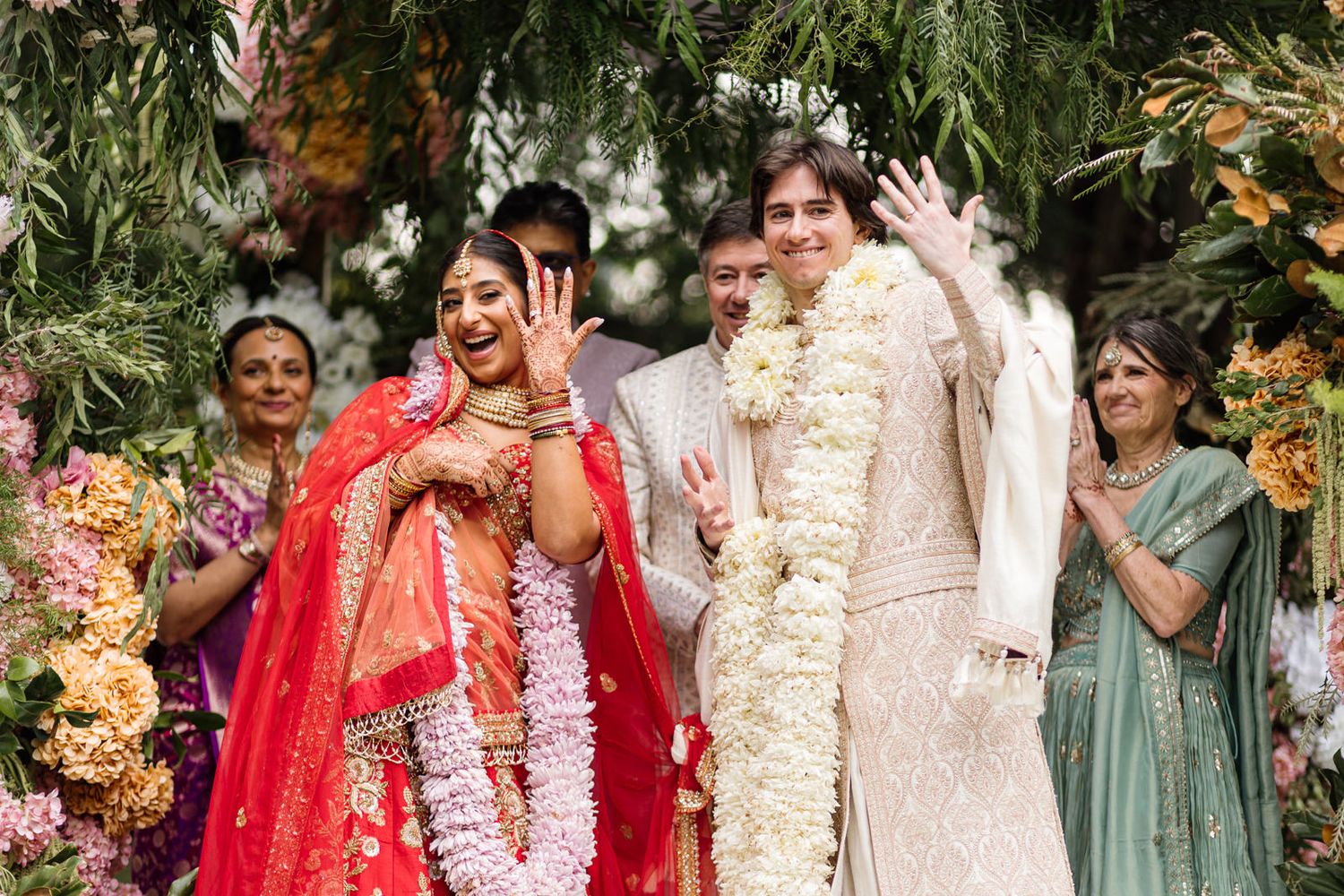 Joyful Indian wedding couple in traditional attire with floral garlands, waving and smiling under lush floral arch.