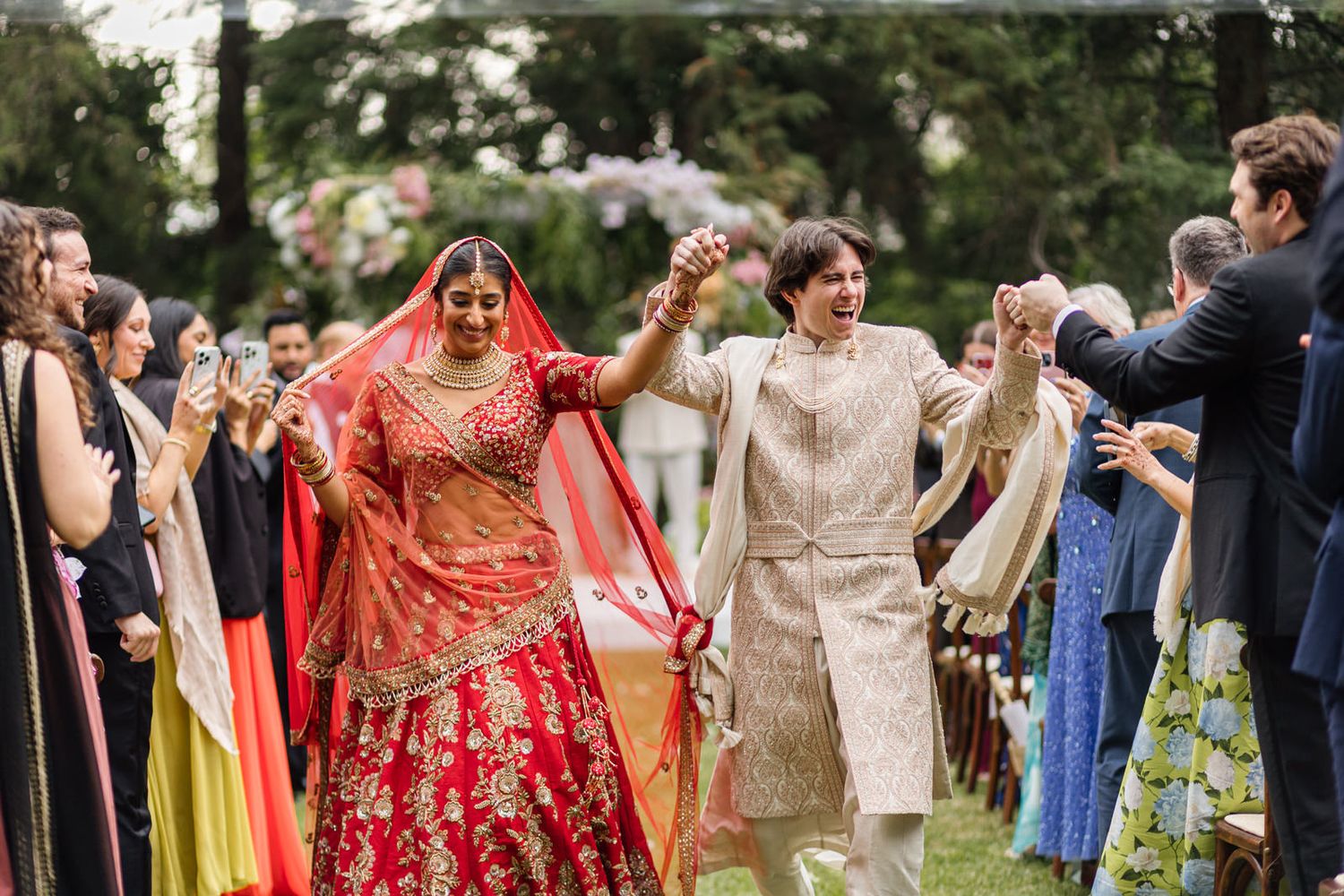 Indian bride in red lehenga and groom in beige sherwani celebrate joyfully walking down the aisle at outdoor wedding.