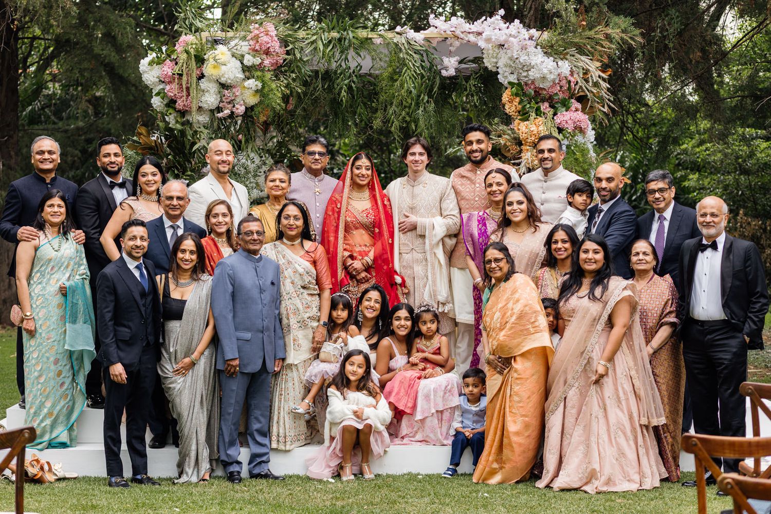 Large Indian wedding family portrait outdoors with floral arch, guests in traditional attire including sari and kurta.
