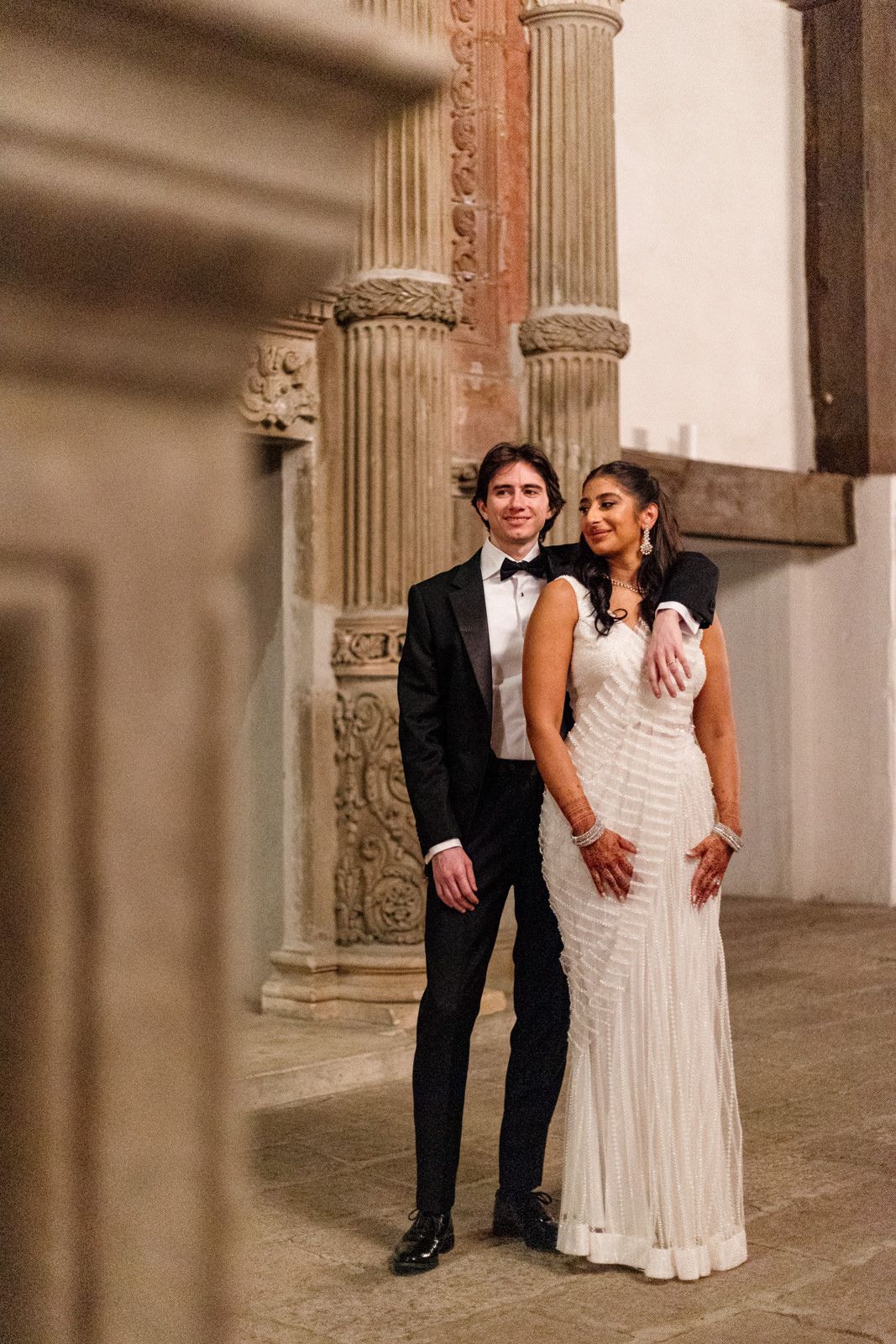 Couple poses elegantly in white beaded gown and black tuxedo amid ornate stone columns.