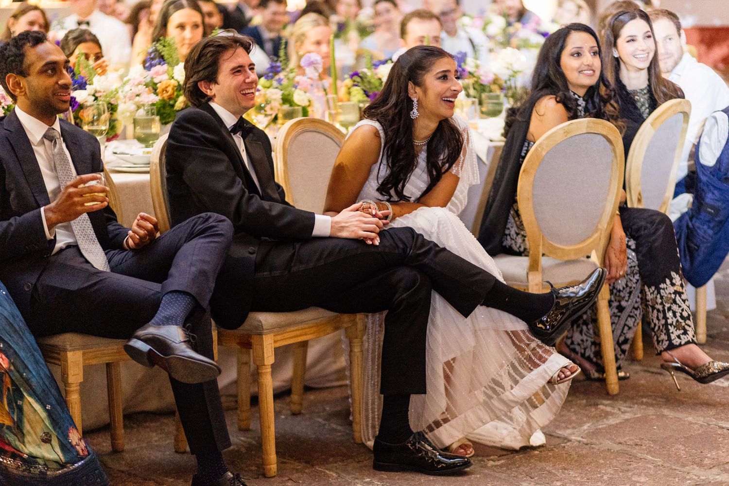 Guests laughing and smiling at a wedding reception, seated in elegant gold chairs at a decorated banquet table.