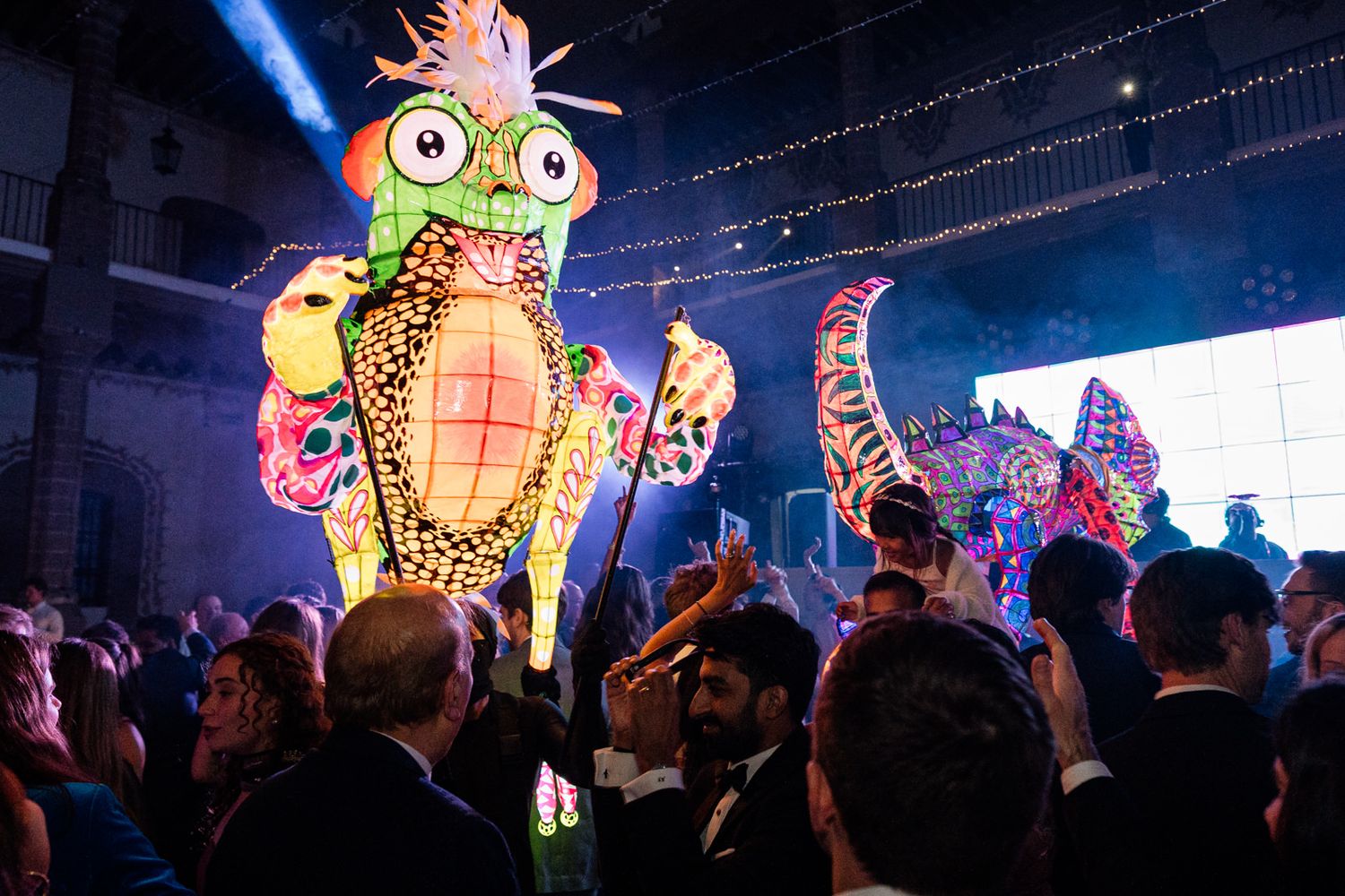 Colorful giant puppet lanterns illuminate a festive indoor party with elegantly dressed guests celebrating.