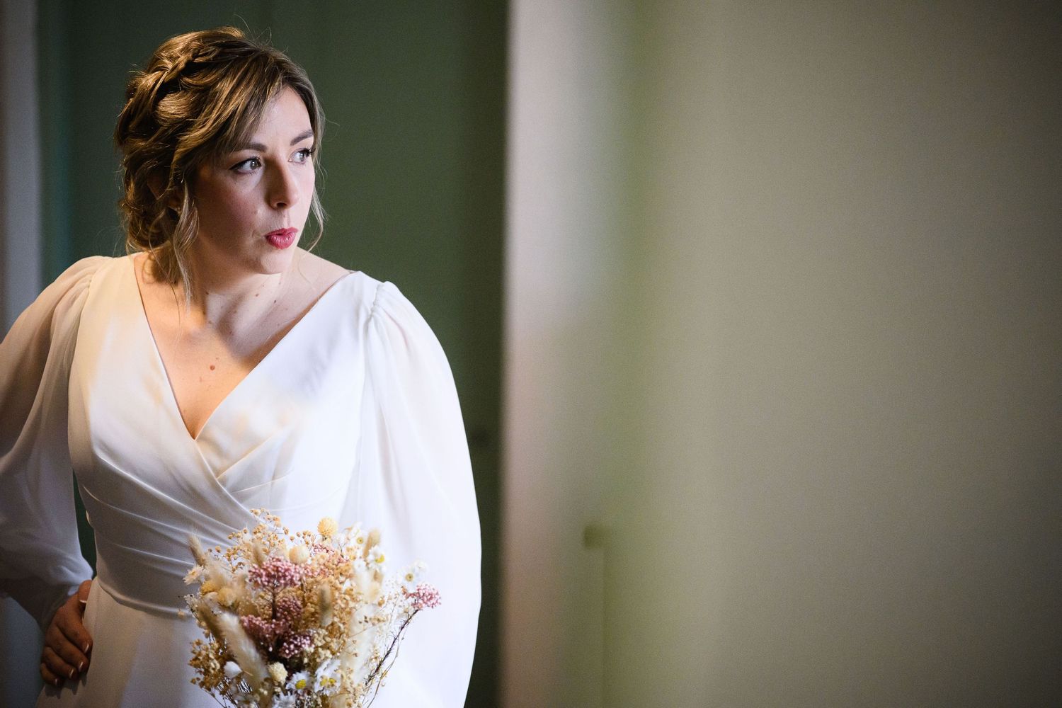 A bride in a classic white wedding dress holds a delicate dried flower bouquet while looking out a window.
